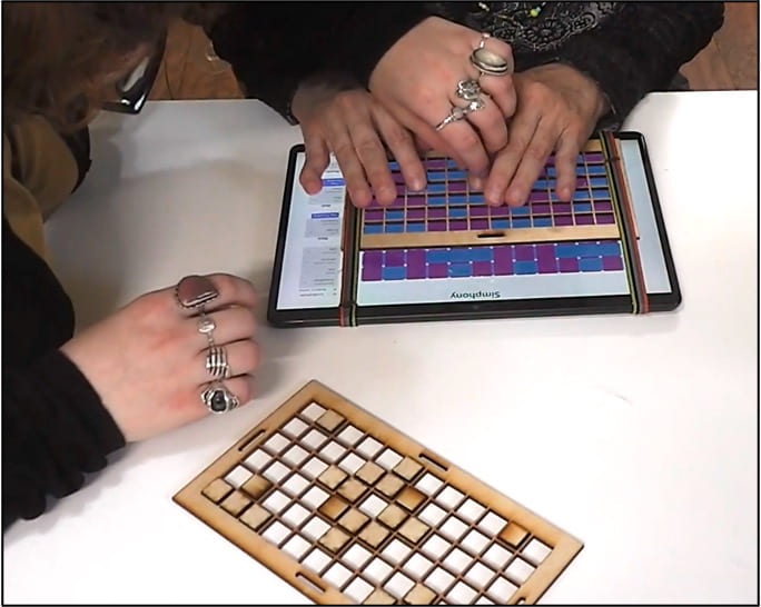 Someone is guiding another person's hand to navigate on the Simphony app with a wooden grid over the tablet. Another wooden grid with blocks arranged in a pattern is placed nearby.