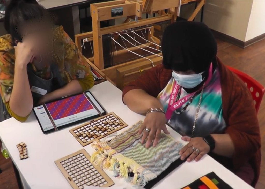 Two people are sitting against a table. One exploring a cloth. On the table are a tablet with the Simphony app open and wooden grid and blocks. Background shows a loom.